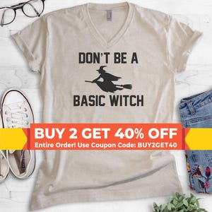 May include: A beige V-neck t-shirt featuring a black graphic of a witch flying on a broomstick with the text DON'T BE A BASIC WITCH printed in black lettering. The shirt is displayed alongside white sneakers and blue denim jeans.