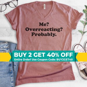 May include: A dusty rose V-neck t-shirt with short sleeves and a heathered texture. The center of the shirt features the black text "Me? Overreacting? Probably." printed in a bold, sans-serif font. The garment is laid flat for display.