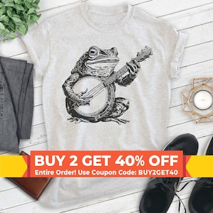 May include: A light heather gray short-sleeve t-shirt featuring a black ink-style illustration of a toad playing a banjo. The graphic is centered on the chest, showcasing the toad in a seated position with its hands on the instrument strings.