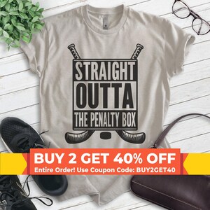May include: A light gray t-shirt with a black graphic design that says "STRAIGHT OUTTA THE PENALTY BOX" with two hockey sticks crossed behind the text. The graphic is in a white box.  The t-shirt has a buy two get 40% off sale with the coupon code BUY2GET40.