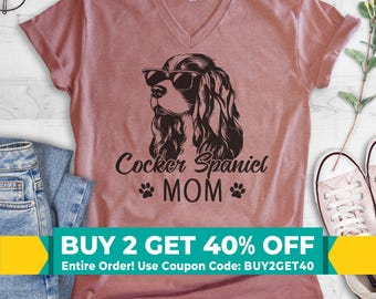 Pode incluir: Uma t-shirt cor de malva com decote em V, apresentando um gráfico preto de um cão Cocker Spaniel a usar óculos de sol. Abaixo do cão, o texto diz Cocker Spaniel MOM, ladeado por dois pequenos ícones de pegadas. Manga curta.