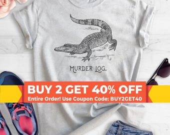 Puede incluir: Una camiseta gris con una ilustración en blanco y negro de un caimán y el texto "MURDER LOG." La camiseta tiene una oferta de compra dos y obtén un 40% de descuento con el código de cupón BUY2GET40.