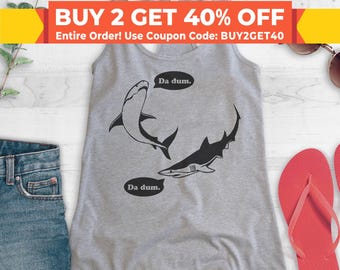 May include: Gray tank top with a graphic of two sharks and the text "Da dum." repeated twice. The tank top is on a white wooden surface.