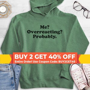 May include: A heather green hooded sweatshirt featuring the black text Me? Overreacting? Probably. centered on the chest. The garment includes a hood with drawstrings and a front kangaroo pocket, displayed with blue denim jeans and white sneakers.