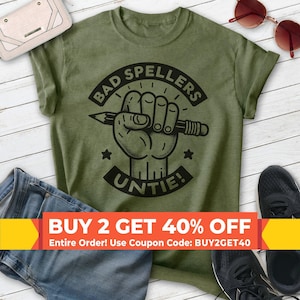 May include: An olive green short-sleeve t-shirt featuring a black graphic of a fist holding a pencil. The text reads BAD SPELLERS UNTIE! The design includes two small stars and circular lines surrounding the central fist and pencil illustration.