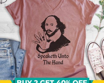May include: A dusty rose V-neck t-shirt featuring a black graphic illustration of William Shakespeare holding up an open hand. Below the illustration, the text reads, "Speaketh Unto The Hand" in a dark, stylized serif font.