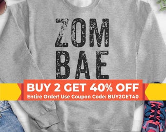 May include: A heather gray crewneck sweatshirt featuring the black distressed text ZOM BAE centered on the chest. The garment has long sleeves and a ribbed neckline, cuffs, and hem. The design is printed in a bold, sans-serif font style.