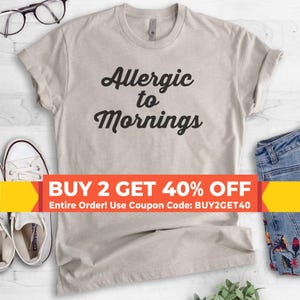 May include: A light beige t-shirt with the words "Allergic to Mornings" in black script. The shirt is displayed with a pair of white sneakers, glasses, and denim jeans.