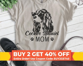 Puede incluir: Una camiseta gris claro con una ilustración en blanco y negro de un cocker spaniel con gafas de sol. El texto "Cocker Spaniel MOM" está impreso debajo de la ilustración. El texto "BUY 2 GET 40% OFF" está impreso en rojo sobre un fondo amarillo en la parte inferior de la imagen.