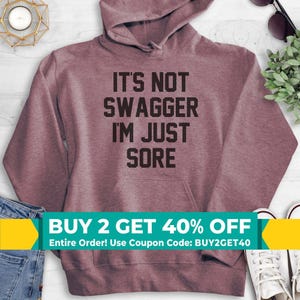 May include: A heathered mauve hooded sweatshirt featuring a front kangaroo pocket and long sleeves. The center of the chest displays the black text IT'S NOT SWAGGER I'M JUST SORE in a bold, sans-serif font. The garment is made of a soft fabric.