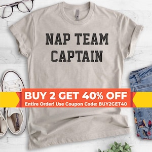 May include: A light beige short-sleeved t-shirt featuring the text NAP TEAM CAPTAIN in bold black block letters centered on the chest. The shirt is displayed alongside a pair of white canvas sneakers and blue denim jeans with floral embroidery.