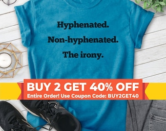 May include: A bright blue short-sleeved t-shirt featuring the black text "Hyphenated. Non-hyphenated. The irony." printed on the front. The shirt has a crew neckline and is displayed flat with a pair of black athletic sneakers and glasses.