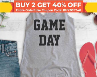 May include: Gray tank top with the text "GAME DAY" in black block letters. The tank top is on a white wooden surface with a pair of red flip flops and a pair of blue denim shorts.