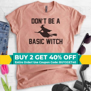 May include: A dusty rose short-sleeved t-shirt featuring a black graphic of a witch flying on a broomstick. The text printed in black reads DON'T BE A BASIC WITCH. The shirt has a crew neckline and is made of a soft, heathered fabric material.