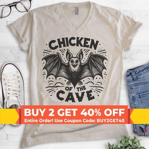 May include: A beige V-neck t-shirt featuring a black graphic of a bat with wings spread wide. The text "CHICKEN OF THE CAVE" is printed in a bold, arched font above and below the bat illustration. The design includes decorative line accents.