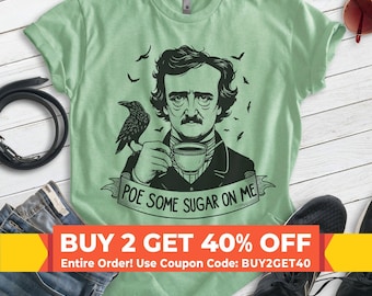 May include: A light green t-shirt featuring a black graphic illustration of Edgar Allan Poe holding a teacup with a raven perched on his shoulder. Below the illustration, a banner displays the text, "POE SOME SUGAR ON ME."