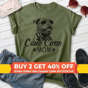 Può includere: Una t-shirt verde oliva con una grafica nera di un cane Cane Corso che indossa occhiali da sole. Sotto il cane, il testo recita Cane Corso MOM con un'icona di impronta di zampa su ogni lato. La maglietta ha maniche corte e girocollo.