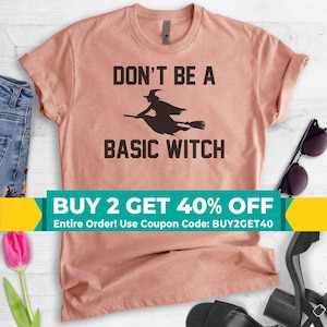 May include: A dusty rose short-sleeved t-shirt featuring a black graphic of a witch flying on a broomstick. The text printed in black reads DON'T BE A BASIC WITCH. The shirt has a crew neckline and a standard fit, presented as a flat lay garment.