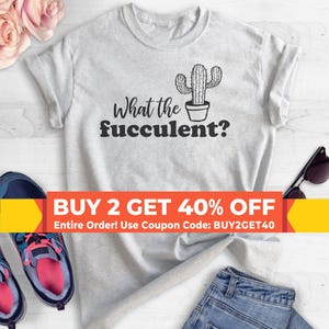 May include: A light gray short-sleeve t-shirt featuring a black graphic of a potted cactus and the text What the fucculent? The shirt is displayed with blue jeans, athletic sneakers, and pink flowers, with a promotional banner across the center.