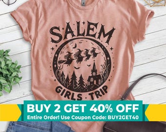 May include: A dusty rose t-shirt featuring a black graphic of witches flying on broomsticks over a forest and a church, with the text SALEM GIRLS TRIP. The design is circular and includes small bats and stars surrounding the central scene.