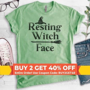 May include: A light green short-sleeve t-shirt featuring a dark green graphic print of a witch hat and a broomstick above the text "Resting Witch Face". The shirt has a crew neckline and is displayed flat with a pair of glasses and denim fabric.