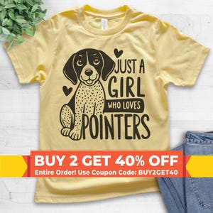 May include: A pale yellow short-sleeve t-shirt featuring a black graphic of a spotted dog and the text "JUST A GIRL WHO LOVES POINTERS" with two small hearts. The shirt has a crew neckline and is displayed flat with a portion of blue denim visible.