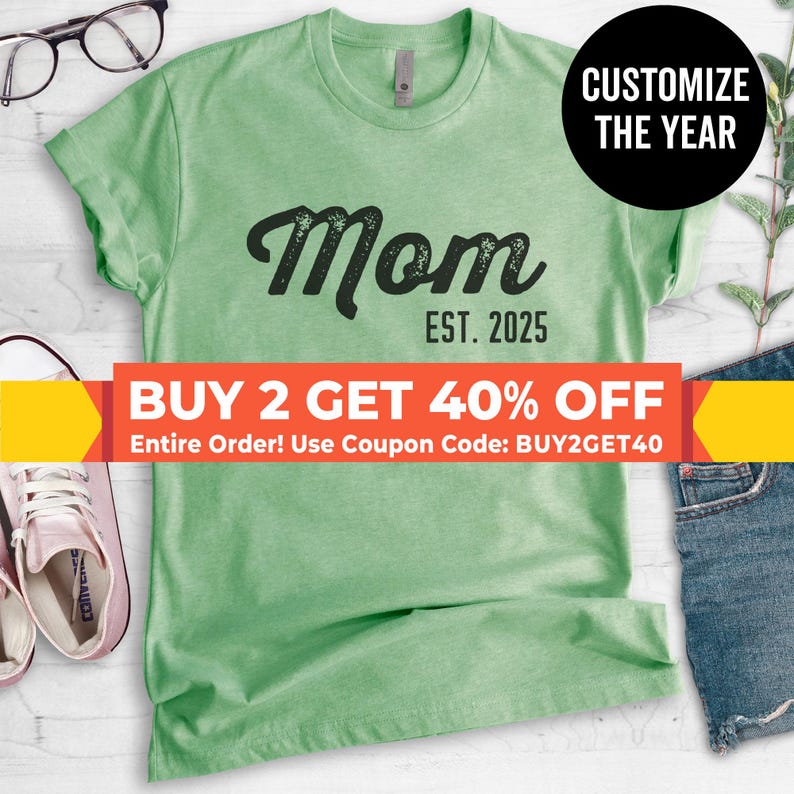 Peut inclure: Un t-shirt vert avec le mot "Mom" en police noire us&eacute;e et le texte "EST. 2025" en dessous. Le t-shirt est en vente avec une offre d'achat de deux pour 40% de r&eacute;duction avec le code promo BUY2GET40.
