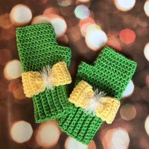 May include: A pair of green crocheted fingerless gloves with yellow bow accents. The gloves are made of a textured yarn and feature a ribbed cuff. The bows are made of a lighter yellow yarn and have a fluffy white center.