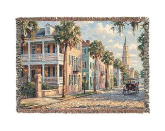 May include: Woven tapestry throw blanket featuring a scenic street view with colorful buildings, palm trees, and a horse-drawn carriage. The artwork depicts a sunny day with a church steeple in the distance. The blanket has a fringed edge.