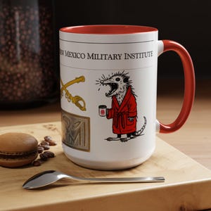 Op de afbeelding: Witte keramische mok met een rode binnenkant en handvat. De mok heeft de tekst "NEW MEXICO MILITARY INSTITUTE" en een cartoon opossum in een rode badjas met een koffiekopje. Een gouden zwaard en embleem staan ook op de mok.