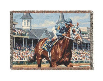 May include: Woven throw blanket featuring a horse race scene. The image depicts a jockey in blue and white riding a brown horse, with a crowd in the background. The blanket has a fringed edge and a detailed, colorful design.