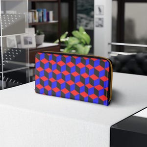Peut inclure: Un portefeuille rectangulaire avec un motif de cubes 3D répété en rouge, bleu et noir. Le portefeuille a une fermeture éclair dorée et est fait d'un matériau lisse. Le portefeuille est posé sur une surface blanche.