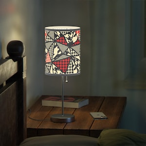 May include: A contemporary table lamp with a patterned lampshade. The shade displays red, black, and cream-coloured triangles. The lamp has a silver metal base and stem. The lamp is on a wooden bedside table.