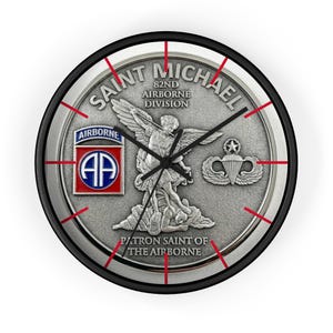 May include: Round wall clock with a black frame and a silver-colored face. The clock face features the words "Saint Michael 82nd Airborne Division" and an image of an angel. Red lines mark the hours.