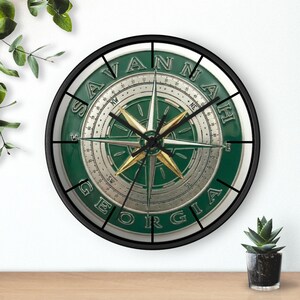 May include: Round wall clock with a black frame, featuring a compass design in green and silver. The clock face displays the text "SAVANNAH GEORGIA" in a circular pattern. The compass needle is gold.