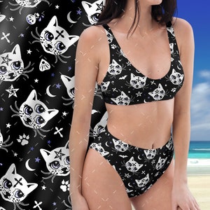 Op de afbeelding: Zwarte tweedelige bikiniset met een herhalend patroon van witte kattengezichten, sterren, manen, kruisen en pootafdrukken. De bikinitop heeft een ronde hals en de broek is hoog getailleerd. De tekst "MOOSE DISCO" is verticaal gedrukt.