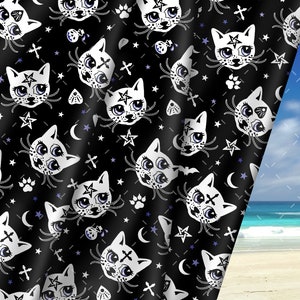 Op de afbeelding: Zwarte stof met een herhalend patroon van witte cartoonkatten met pentagrammen op hun voorhoofd, sterren, manen, kruisen en potafdrukken. De stof is gedrapeerd over een strandscherm.