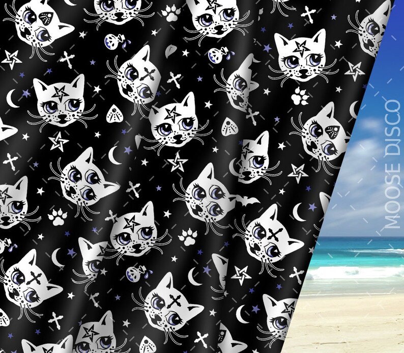 Op de afbeelding: Zwarte stof met een herhalend patroon van witte cartoonkatten met pentagrammen op hun voorhoofd, sterren, manen, kruisen en potafdrukken. De stof is gedrapeerd over een strandscherm.