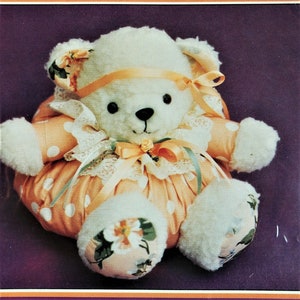 May include: A white teddy bear sewing pattern with the title "Q.T. Bunz A bear sewing pattern". The bear is wearing a peach-colored dress with white polka dots and a white lace trim. The bear has a yellow ribbon around its neck and a pink rose on its chest.