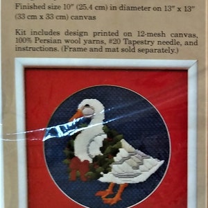May include: A framed needlepoint design of a white duck with a red holly wreath around its neck. The duck is standing on a blue background with a red border. The finished size is 10 inches in diameter on a 13 inch by 13 inch canvas.