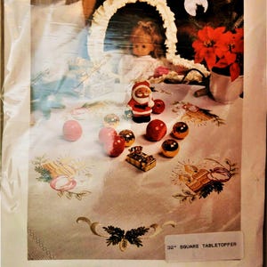 May include: A white tablecloth with a Christmas apple embroidery design. The tablecloth is 32 inches square and is pre-stamped for hand embroidery. The image also shows a doll, a Santa Claus figurine, and red and gold ornaments.