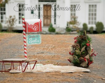 May include: A red mailbox with a white snow cap and a sign that says "Letters to Santa" is on a red and white striped pole. A small Christmas tree with pine cones and red berries is in the foreground. A red sled is on the ground.