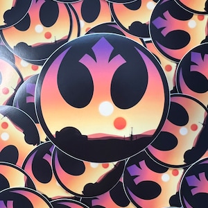 May include: A stack of round stickers featuring a black Rebel Alliance symbol with a sunset background and two suns. The stickers are printed on white paper.