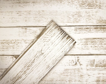 May include: A close-up of a white painted wood plank with a distressed finish. The wood has a light brown undertone and shows signs of wear and tear.