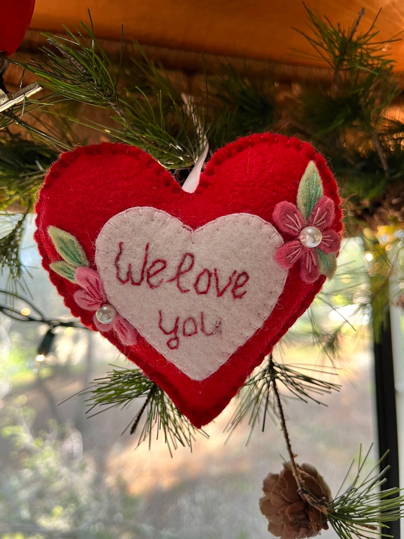 Może przedstawiać: Czerwona filcowa ozdoba w kształcie serca z białym sercem pośrodku, wyszytym napisem "We love you". Dołączone są r&oacute;żowe kwiaty z tkaniny i zielone liście. Biała wstążka znajduje się na g&oacute;rze do zawieszenia. Ozdoba wisi na choince.