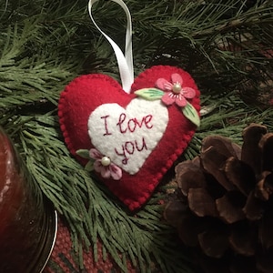 Może przedstawiać: Czerwona filcowa ozdoba w kształcie serca z napisem "I love you" wyhaftowanym na białym sercu. Ozdoba jest ozdobiona r&oacute;żowymi kwiatami i zielonymi liśćmi. Do zawieszenia przymocowana jest biała wstążka. Ozdoba otoczona jest zielenią i szyszką.