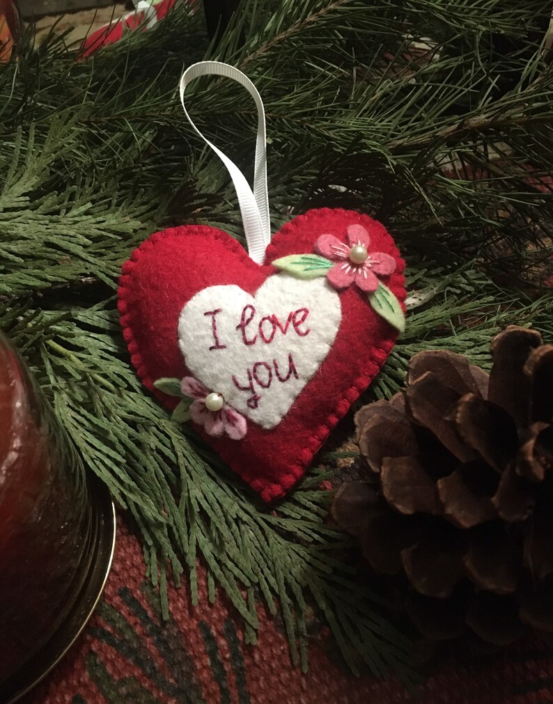 Może przedstawiać: Czerwona filcowa ozdoba w kształcie serca z napisem "I love you" wyhaftowanym na białym sercu. Ozdoba jest ozdobiona r&oacute;żowymi kwiatami i zielonymi liśćmi. Do zawieszenia przymocowana jest biała wstążka. Ozdoba otoczona jest zielenią i szyszką.