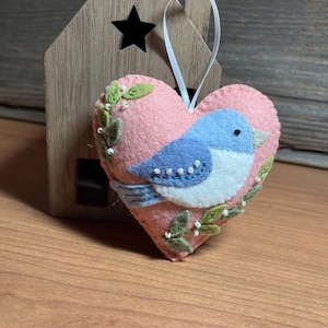 May include: A pink felt heart-shaped ornament with a blue and white bird design. The bird is surrounded by green leaves and white beads. A white ribbon is attached for hanging. A wooden house-shaped decoration is in the background.