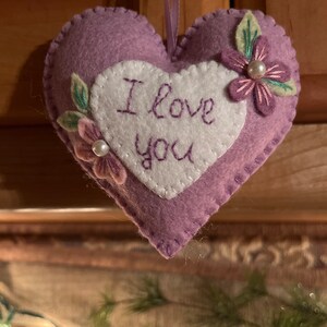 Może przedstawiać: Lawendowa filcowa ozdoba w kształcie serca z białym sercem pośrodku. Na białym sercu wyhaftowano fioletową nicią słowa "I love you". Ozdoba jest ozdobiona filcowymi kwiatami i liśćmi oraz perłą. Do zawieszenia przymocowana jest fioletowa wstążka.