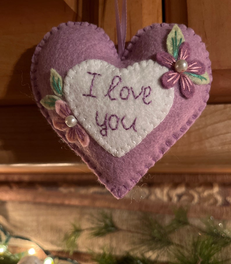 Może przedstawiać: Lawendowa filcowa ozdoba w kształcie serca z białym sercem pośrodku. Na białym sercu wyhaftowano fioletową nicią słowa "I love you". Ozdoba jest ozdobiona filcowymi kwiatami i liśćmi oraz perłą. Do zawieszenia przymocowana jest fioletowa wstążka.